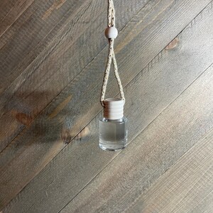 K&ouml;nnte beinhalten: Eine klare Glasflasche mit einem Holzverschluss, die an einem beigefarbenen Seil mit Holzperlen h&auml;ngt. Die Flasche ist mit einer klaren Fl&uuml;ssigkeit gef&uuml;llt.