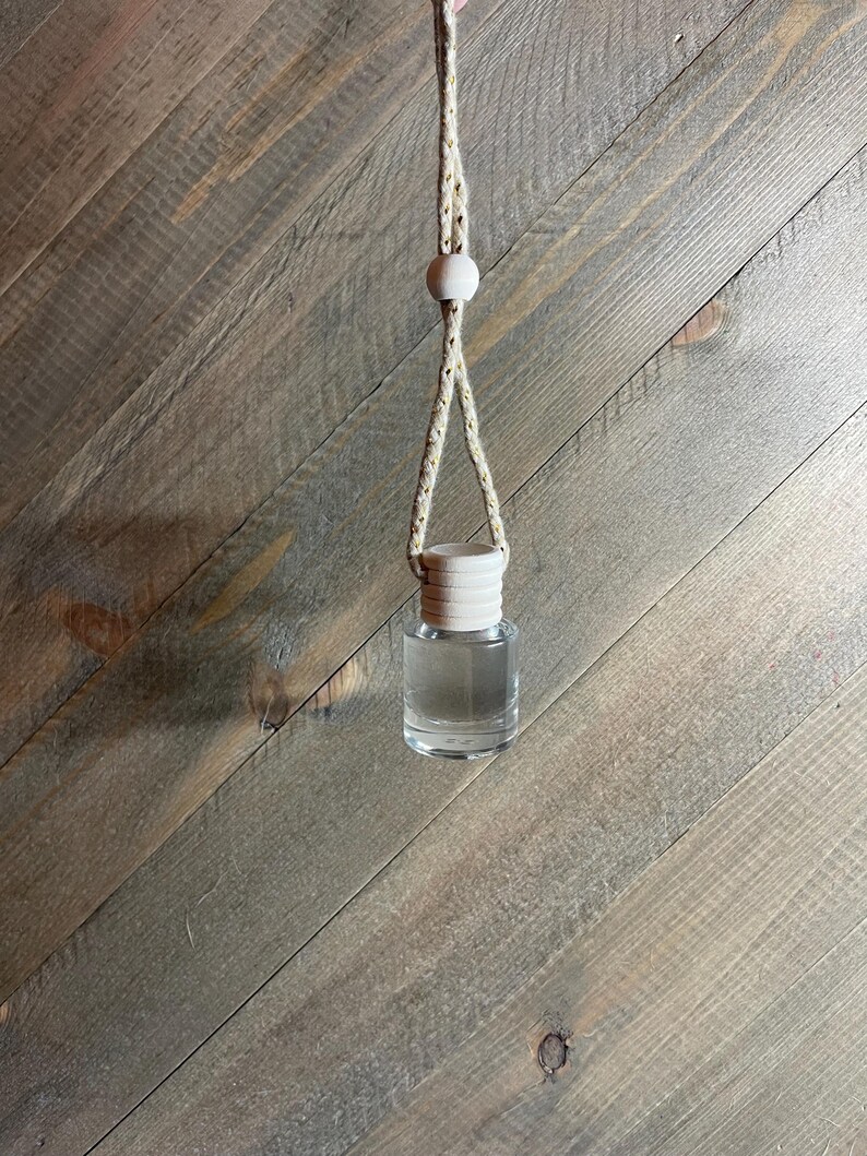 K&ouml;nnte beinhalten: Eine klare Glasflasche mit einem Holzverschluss, die an einem beigefarbenen Seil mit Holzperlen h&auml;ngt. Die Flasche ist mit einer klaren Fl&uuml;ssigkeit gef&uuml;llt.