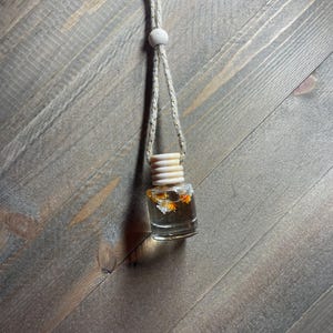 K&ouml;nnte beinhalten: Eine klare Glasflasche mit einem Holzverschluss und einer daran befestigten Schnur. Die Flasche enth&auml;lt getrocknete Blumen und ist mit einer klaren Fl&uuml;ssigkeit gef&uuml;llt.