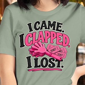 May include: A sage green t-shirt featuring the black text I CAME. I CLAPPED. I LOST. surrounding a pink hand-shaped clapper and three pink tickets. The design uses bold lettering and a pink underline beneath the final word.