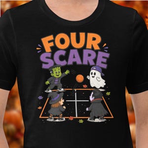 May include: A black t-shirt featuring a graphic of a Frankenstein monster, a ghost, a witch, and a vampire playing four square. The text "FOUR SCARE" is printed in orange and purple above the characters, with small autumn leaves scattered around.