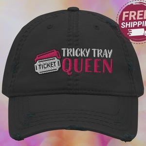 May include: Black distressed baseball cap with the embroidered text "TRICKY TRAY QUEEN" in white and pink. A pink and white ticket graphic is on the left side of the cap. The cap has a curved brim and a distressed look.