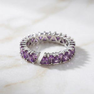 Puede incluir: Un anillo plateado con una fila de piedras preciosas triangulares moradas. El anillo tiene un diseño abierto, con un hueco en la banda. Las piedras están engastadas muy juntas, creando una línea continua de color. El anillo se muestra sobre una superficie blanca.