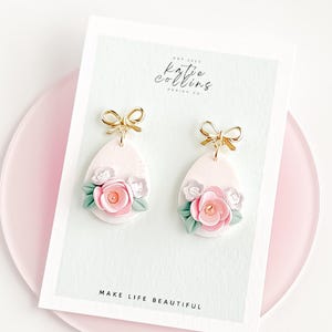Könnte beinhalten: Hellrosa tropfenförmige Ohrringe mit goldenen Schleifen. Jeder Ohrring hat rosa und weiße Blumen mit grünen Blättern. Sie werden auf einer weißen Karte mit dem Text "MAKE LIFE BEAUTIFUL" präsentiert.