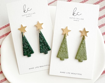 May include: Two pairs of Christmas tree earrings. One pair is dark green, the other is olive green. Each earring has a gold star at the top and is attached to a white card with the text "Katie Collins Design Co." and "Make Life Beautiful."