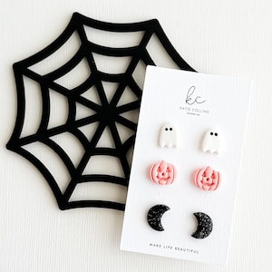 May include: A black spiderweb decoration and a card with Halloween-themed earrings. The earrings include white ghosts, pink jack-o'-lanterns, and black crescent moons. The card has the text "MAKE LIFE BEAUTIFUL".