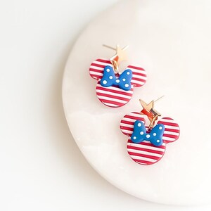 Puede incluir: Un par de pendientes a rayas rojas y blancas con forma de cabeza de rat&oacute;n con un lazo azul y detalles de estrella dorada. Los pendientes est&aacute;n sobre una superficie blanca.