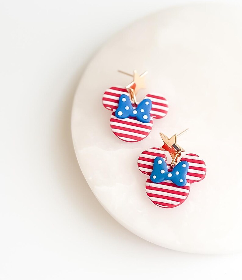 Puede incluir: Un par de pendientes a rayas rojas y blancas con forma de cabeza de rat&oacute;n con un lazo azul y detalles de estrella dorada. Los pendientes est&aacute;n sobre una superficie blanca.