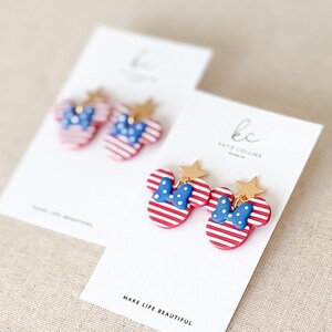 Puede incluir: Pendientes a rayas rojas y blancas con forma de cabeza de rat&oacute;n, con lazo azul y detalles de estrella dorada. Los pendientes se muestran en tarjetas blancas con el texto "MAKE LIFE BEAUTIFUL".