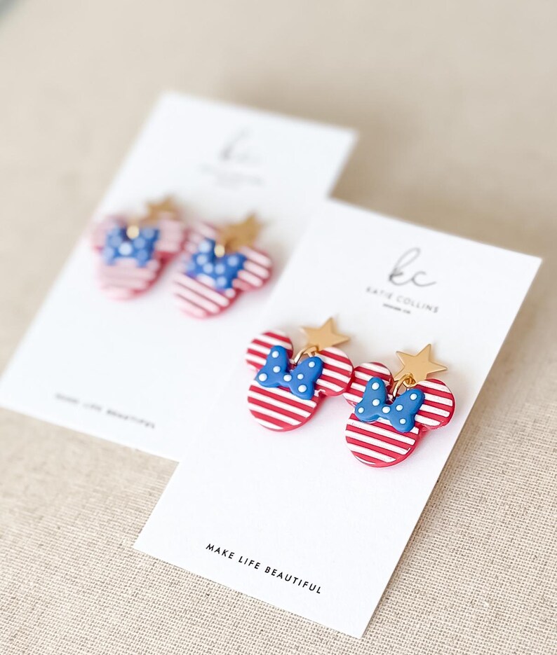 Puede incluir: Pendientes a rayas rojas y blancas con forma de cabeza de rat&oacute;n, con lazo azul y detalles de estrella dorada. Los pendientes se muestran en tarjetas blancas con el texto "MAKE LIFE BEAUTIFUL".