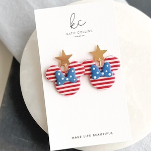 Puede incluir: Pendientes con forma de Minnie Mouse, a rayas rojas y blancas, con lazos azules de lunares y detalles de estrellas doradas. Se presentan en una tarjeta blanca con el texto "MAKE LIFE BEAUTIFUL" y el logotipo del dise&ntilde;ador. Un accesorio divertido.