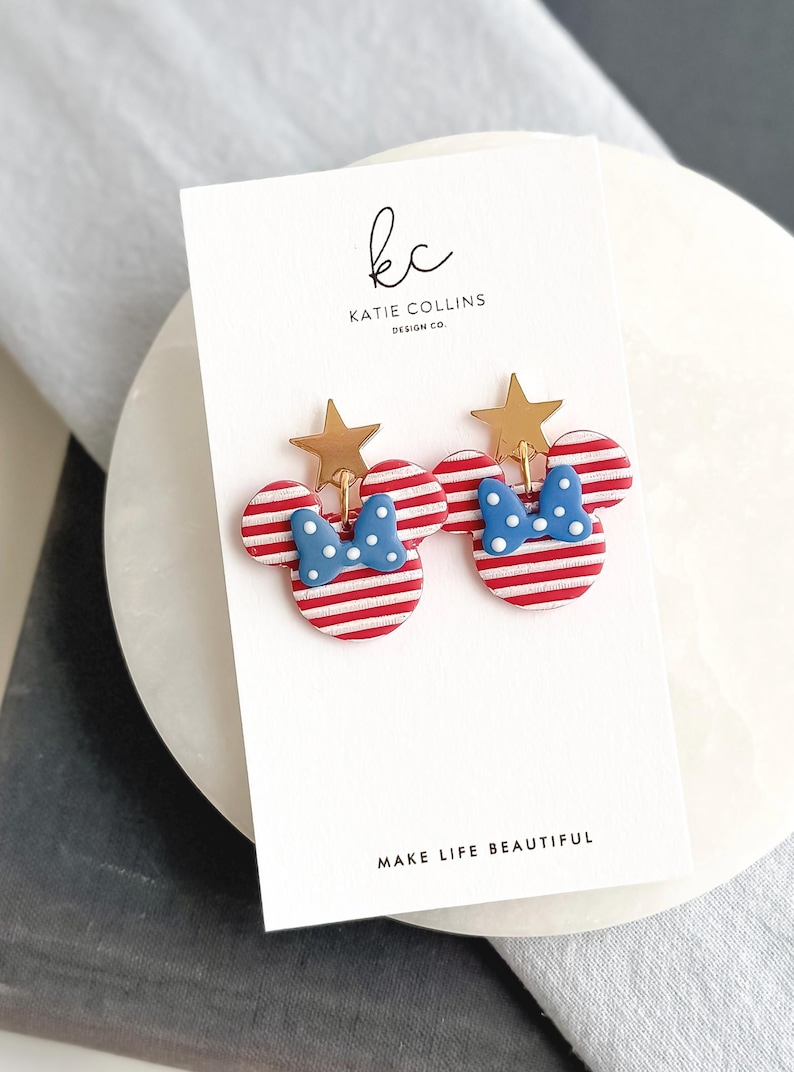 Puede incluir: Pendientes con forma de Minnie Mouse, a rayas rojas y blancas, con lazos azules de lunares y detalles de estrellas doradas. Se presentan en una tarjeta blanca con el texto "MAKE LIFE BEAUTIFUL" y el logotipo del dise&ntilde;ador. Un accesorio divertido.