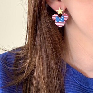 Puede incluir: Pendientes con forma de Minnie Mouse a rayas rojas y blancas, con un lazo azul y una estrella dorada. Un accesorio festivo, perfecto para a&ntilde;adir un toque de diversi&oacute;n a cualquier atuendo.