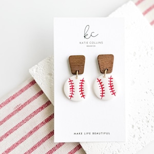 May include: Baseball-themed earrings with white, round pendants featuring red stitching, paired with rectangular wooden tops. The earrings are displayed on a white card with the text "MAKE LIFE BEAUTIFUL".
