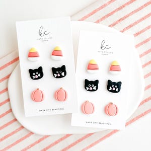 Puede incluir: Dos juegos de pendientes de temática de Halloween en tarjetas blancas. Cada juego incluye un par de pendientes de maíz dulce, gato negro y calabaza. Las tarjetas tienen el texto "kc Katie Collins Design Co. Make Life Beautiful."
