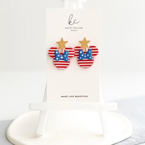 Puede incluir: Un par de pendientes con forma de Mickey Mouse a rayas rojas y blancas con lazos de lunares azules y detalles de estrellas doradas. Los pendientes se exhiben en una tarjeta blanca con el texto "MAKE LIFE BEAUTIFUL" y el logotipo del dise&ntilde;ador.