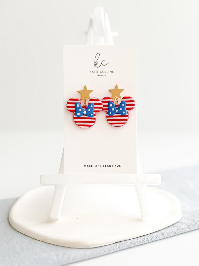 Puede incluir: Un par de pendientes con forma de Mickey Mouse a rayas rojas y blancas con lazos de lunares azules y detalles de estrellas doradas. Los pendientes se exhiben en una tarjeta blanca con el texto "MAKE LIFE BEAUTIFUL" y el logotipo del dise&ntilde;ador.