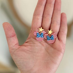 Puede incluir: Un par de pendientes patri&oacute;ticos con un dise&ntilde;o de cabeza de Mickey Mouse. Los pendientes tienen orejas a rayas rojas y blancas, un lazo azul con lunares blancos y una estrella dorada en la parte superior. Los pendientes se sostienen en una mano.