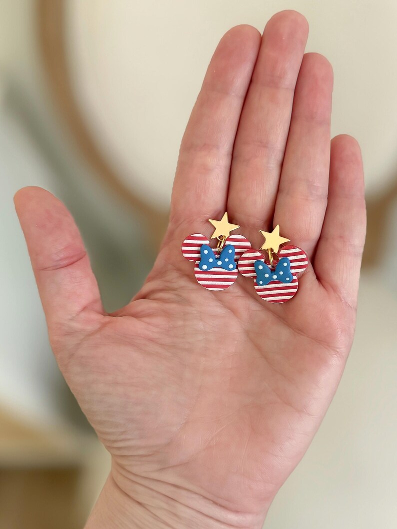 Puede incluir: Un par de pendientes patri&oacute;ticos con un dise&ntilde;o de cabeza de Mickey Mouse. Los pendientes tienen orejas a rayas rojas y blancas, un lazo azul con lunares blancos y una estrella dorada en la parte superior. Los pendientes se sostienen en una mano.