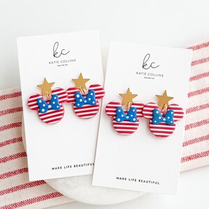 Puede incluir: Dos pares de pendientes con forma de Mickey Mouse a rayas rojas y blancas, con lazos azules de lunares y estrellas doradas. Se exhiben en tarjetas blancas con el texto "MAKE LIFE BEAUTIFUL" y el logotipo del dise&ntilde;ador.