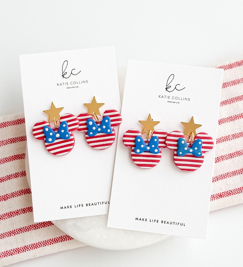 Puede incluir: Dos pares de pendientes con forma de Mickey Mouse a rayas rojas y blancas, con lazos azules de lunares y estrellas doradas. Se exhiben en tarjetas blancas con el texto "MAKE LIFE BEAUTIFUL" y el logotipo del dise&ntilde;ador.