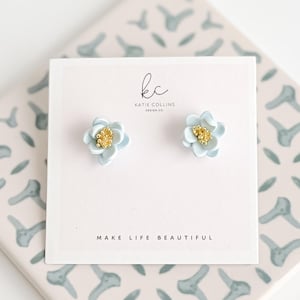 May include: A pair of light blue flower stud earrings with gold centers, displayed on a white card. The card features the text "MAKE LIFE BEAUTIFUL" and the designer's logo. The earrings are set on a patterned surface.
