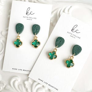 May include: A pair of green and gold earrings with a clover-shaped charm. The earrings are displayed on a white card with the text "kc KATIE COLLINS DESIGN CO. MAKE LIFE BEAUTIFUL".