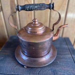 May include: A vintage copper kettle with a wooden handle and a copper stand. The kettle has a tarnished finish and is in good condition.