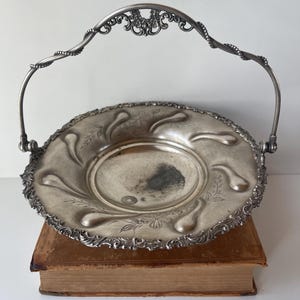 Pode incluir: Travessa de servir em metal prateado com pega decorativa. A travessa tem uma borda ornamentada com motivos florais e uma área central ligeiramente escurecida. A pega é arqueada e apresenta floreios decorativos. A travessa está sobre um livro antigo.