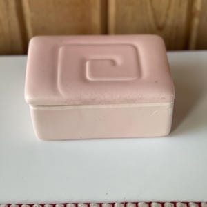 May include: A small, pink ceramic trinket box with a swirl design on the lid. The box has a white interior.
