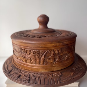 May include: A round, wooden box with a lid and a base, all intricately carved with a detailed relief design. The box is a warm brown color, and the lid has a small, rounded handle. The base is a wide, flat circle.
