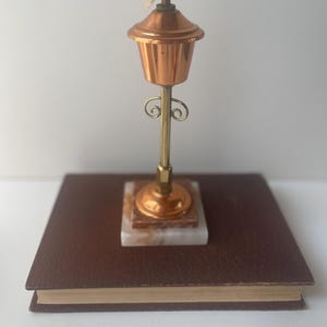 Puede incluir: Una pequeña lámpara de aceite de cobre con un tallo de latón y una base de mármol. La lámpara tiene un diseño decorativo y una mecha. La lámpara está colocada sobre un libro marrón.