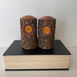 May include: Two cylindrical salt and pepper shakers crafted from natural wood logs with rough, textured bark exteriors. Each shaker features a smooth, sanded oval inlay on the front with the words Salt and Pepper written in black script.