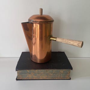 May include: A polished copper coffee pot with a cylindrical body, a wide spout, and a lid featuring a small wooden knob. A long, straight wooden handle is attached to the side with a brass-colored metal bracket, resting on a thick, dark-bound book.