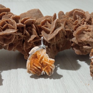 Puede incluir: Tres colgantes de rosa del desierto con alambre de plata, expuestos frente a una formación más grande de rosa del desierto. Los colgantes son de varios tonos de naranja y marrón, mostrando las formaciones cristalinas naturales.