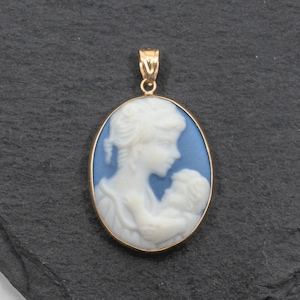 May include: An oval-shaped pendant featuring a white relief carving of a person holding a child against a light blue background. The cameo is encased in a polished gold-toned metal frame with a matching gold-toned bail at the top for hanging.