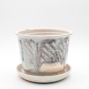 May include: A ceramic plant pot with a matching saucer. The pot is cylindrical with a wide opening and features a glaze with shades of blue, purple, and cream. The saucer is round and has a slightly raised edge. The pot and saucer are suitable for indoor or outdoor use.