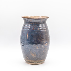 May include: A ceramic vase with a glossy, dark blue glaze featuring mottled brown and tan undertones. The vessel has a wide, flared rim, a rounded body that tapers toward the base, and a smooth, reflective surface with visible variations in color.