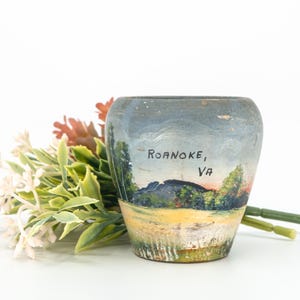 May include: A small, rounded ceramic planter featuring a hand-painted landscape scene of a mountain range and trees. The text "ROANOKE, VA" is clearly printed in black on the front. The base is painted in shades of yellow and green.