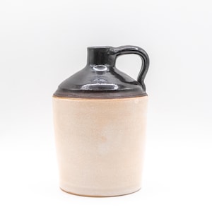 May include: A ceramic jug featuring a cylindrical body with a matte, light beige finish and a contrasting glossy, dark brown glazed neck and handle. The vessel has a narrow opening at the top and a single curved handle attached to the side.