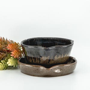 May include: A dark brown and black glazed ceramic bowl with a matching saucer, featuring a small pouring spout on the rim. The bowl has a glossy finish with visible drip patterns, and a small cluster of artificial orange and green foliage sits beside it.