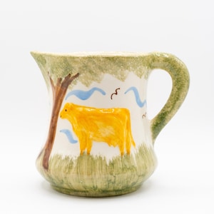 May include: A ceramic pitcher with a hand-painted design. The pitcher features a yellow cow, a brown tree, blue wavy lines, and green grass and foliage. The handle and rim are green.