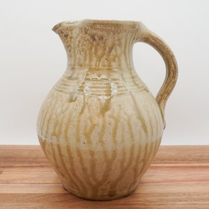 May include: A ceramic pitcher with a light brown glaze and a textured surface. The pitcher has a wide base and a narrow neck with a handle.