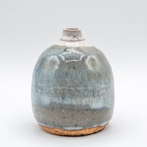 May include: A small, handcrafted ceramic vase with a rounded body and a narrow neck. The vase features a gradient of colors, transitioning from a light blue at the top to a darker gray-brown at the base. The base has a textured, natural finish.