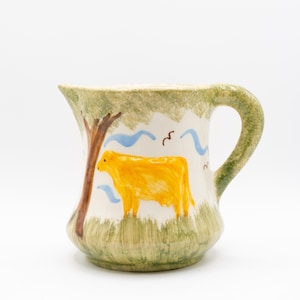 May include: A ceramic pitcher with a hand-painted design. The pitcher features a yellow cow, a brown tree, and blue wavy lines against a white background. The handle and rim are green, and the pitcher has a rustic, farmhouse aesthetic.