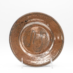 May include: A round, brown ceramic plate with a textured surface and a decorative swirl pattern in the center. The plate has a slightly raised rim and is displayed on a clear stand. The plate's color is a warm, earthy tone.