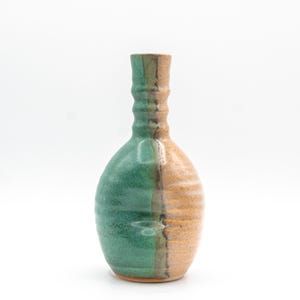 May include: A ceramic vase with a bulbous body and a narrow, ribbed neck. The surface features a distinct vertical split, with one side finished in a textured matte green glaze and the other side in a speckled, earthy tan glaze with horizontal ridges.