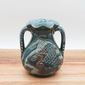 May include: A green ceramic vase with two handles and a fish design. The vase has a textured surface and a scalloped rim.