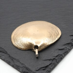 May include: A gold-toned pendant shaped like a clam shell with fine, radiating ridges across its surface. The pendant features a small, integrated metal loop at the base for attachment, showcasing a smooth, metallic finish with subtle texture.