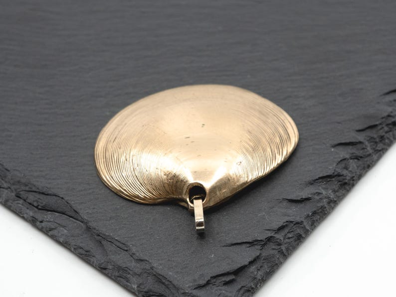 May include: A gold-toned pendant shaped like a clam shell with fine, radiating ridges across its surface. The pendant features a small, integrated metal loop at the base for attachment, showcasing a smooth, metallic finish with subtle texture.